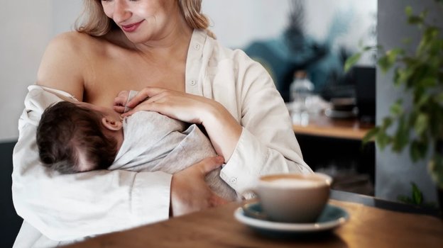 Женщина кормит грудью ребенка и пьет кофе.