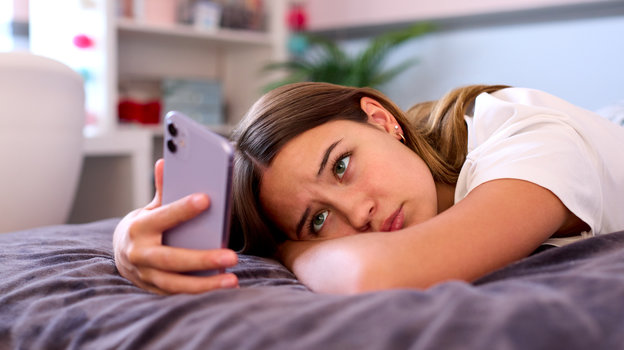 Грустная девушка лежит на кровати и смотрит в телефон